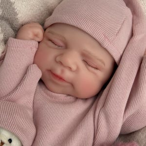 Pu&ograve; includere: Una bambola neonato realistica con occhi chiusi ed espressione serena, che indossa una maglia a maniche lunghe a coste rosa polvere e un berretto abbinato. La bambola ha la pelle chiara, sopracciglia dipinte e labbra rosa, mani vicino al viso.