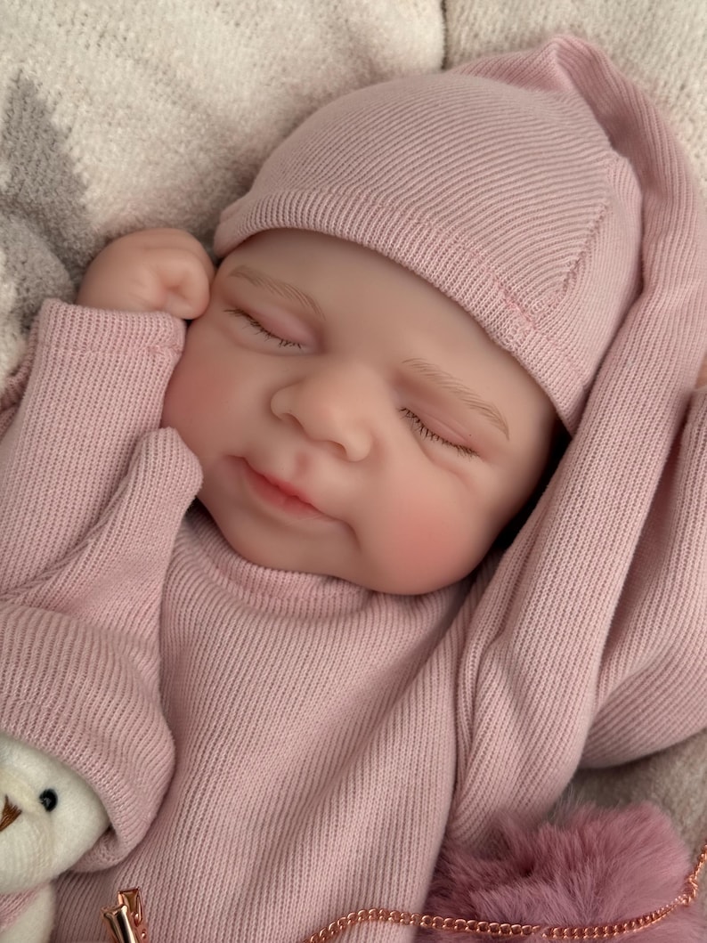 Pu&ograve; includere: Una bambola neonato realistica con occhi chiusi ed espressione serena, che indossa una maglia a maniche lunghe a coste rosa polvere e un berretto abbinato. La bambola ha la pelle chiara, sopracciglia dipinte e labbra rosa, mani vicino al viso.