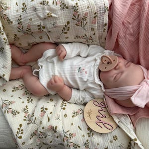 May include: A realistic baby doll wearing a white long-sleeved ribbed bodysuit with a small floral print, a light pink headband with a bow, and a beige dummy. The doll rests on a cream-coloured muslin blanket with a green and pink floral pattern.