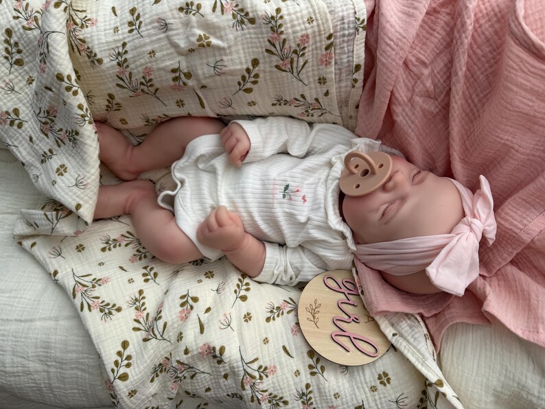 May include: A realistic baby doll wearing a white long-sleeved ribbed bodysuit with a small floral print, a light pink headband with a bow, and a beige dummy. The doll rests on a cream-coloured muslin blanket with a green and pink floral pattern.