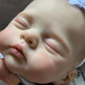 May include: A realistic reborn doll with pale skin, closed eyes, and painted eyebrows. The doll has a small nose, pink lips, and rosy cheeks. A white fabric headband with a small flower sits on the head, and the doll wears a white shirt with a small blue and green floral pattern.