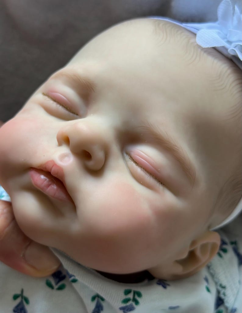 May include: A realistic reborn doll with pale skin, closed eyes, and painted eyebrows. The doll has a small nose, pink lips, and rosy cheeks. A white fabric headband with a small flower sits on the head, and the doll wears a white shirt with a small blue and green floral pattern.
