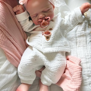 May include: A realistic baby doll with closed eyes, wearing a white textured long-sleeve shirt and matching pants with small brown buttons. The doll has a light pink knotted headband, a metallic rose gold pacifier, and a matching rose gold pendant.