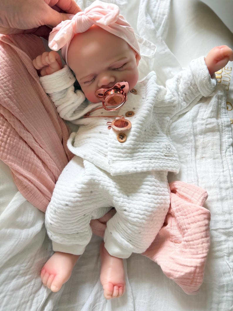 May include: A realistic baby doll with closed eyes, wearing a white textured long-sleeve shirt and matching pants with small brown buttons. The doll has a light pink knotted headband, a metallic rose gold pacifier, and a matching rose gold pendant.