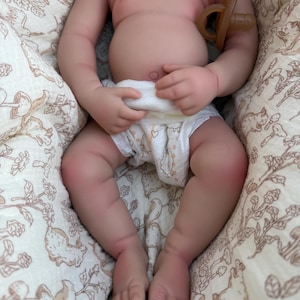 May include: A lifelike baby doll with a pacifier, wearing a diaper with heart designs. The doll is lying on a patterned blanket with a beige background and a design of squirrels and flowers. The doll's skin is a realistic tone.