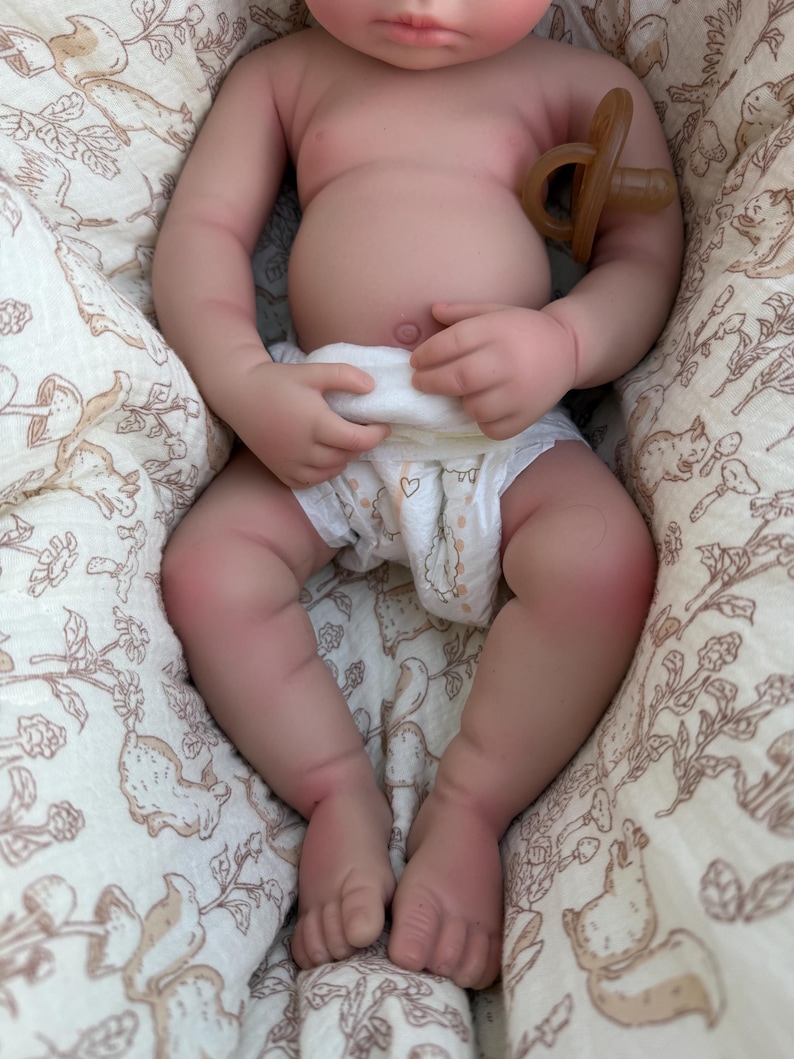 May include: A lifelike baby doll with a pacifier, wearing a diaper with heart designs. The doll is lying on a patterned blanket with a beige background and a design of squirrels and flowers. The doll's skin is a realistic tone.