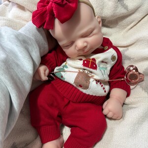 Pode incluir: Uma boneca beb&eacute; a dormir vestida com uma camisola de malha vermelha com um gr&aacute;fico de rena e presente, cal&ccedil;as de malha vermelhas a combinar e uma fita grande com la&ccedil;o de veludo cotel&ecirc; vermelho. Uma chupeta em ouro rosa est&aacute; presa &agrave; camisola.