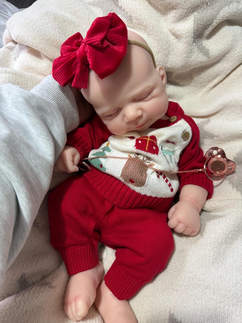 Pode incluir: Uma boneca beb&eacute; a dormir vestida com uma camisola de malha vermelha com um gr&aacute;fico de rena e presente, cal&ccedil;as de malha vermelhas a combinar e uma fita grande com la&ccedil;o de veludo cotel&ecirc; vermelho. Uma chupeta em ouro rosa est&aacute; presa &agrave; camisola.