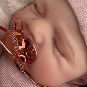 Pu&ograve; includere: Una bambola realistica con pelle chiara, occhi chiusi e ciglia castano chiaro, che indossa un berretto a coste rosa tenue e una maglietta abbinata. Un ciuccio in oro rosa metallizzato con un centro circolare testurizzato &egrave; in bocca, con catenella.