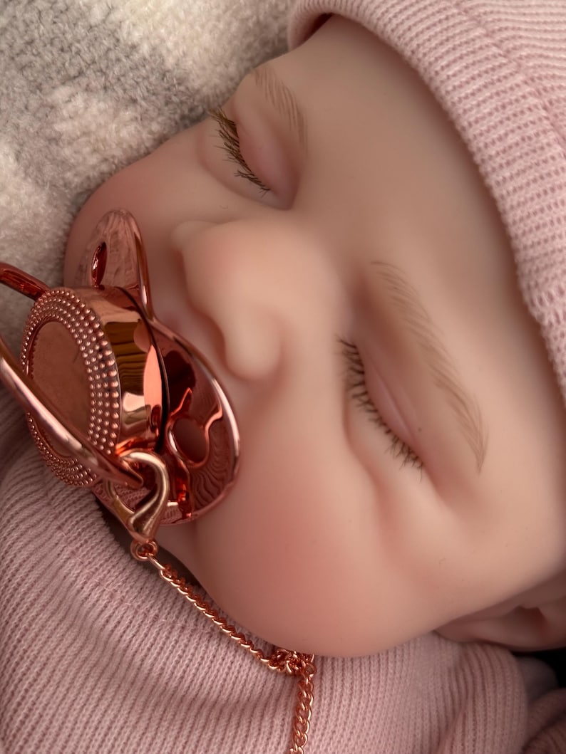 Pu&ograve; includere: Una bambola realistica con pelle chiara, occhi chiusi e ciglia castano chiaro, che indossa un berretto a coste rosa tenue e una maglietta abbinata. Un ciuccio in oro rosa metallizzato con un centro circolare testurizzato &egrave; in bocca, con catenella.