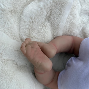 May include: A realistic doll with light skin tone featuring detailed toes and soles of the feet, wearing a plain light purple onesie. The doll is positioned with its legs bent, showcasing the lifelike texture and soft appearance of the limbs.