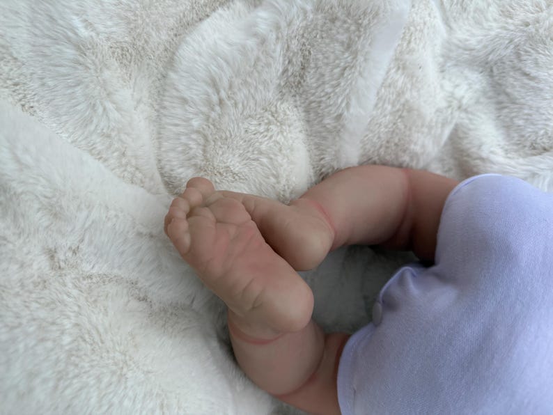 May include: A realistic doll with light skin tone featuring detailed toes and soles of the feet, wearing a plain light purple onesie. The doll is positioned with its legs bent, showcasing the lifelike texture and soft appearance of the limbs.