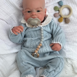 K&ouml;nnte beinhalten: Eine realistische Babypuppe mit Glatze, bekleidet mit einem hellblauen Zopfmuster-Pullover und passender Hose mit wei&szlig;em Saum. Die Puppe tr&auml;gt einen beigen Schnuller an einer grau-beigen Perlenkette und hat einen runden, bunten Silikon-Bei&szlig;ring.
