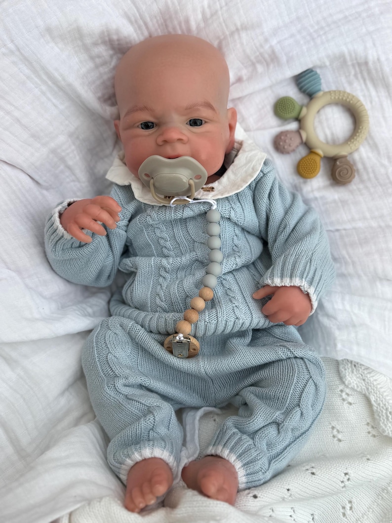 K&ouml;nnte beinhalten: Eine realistische Babypuppe mit Glatze, bekleidet mit einem hellblauen Zopfmuster-Pullover und passender Hose mit wei&szlig;em Saum. Die Puppe tr&auml;gt einen beigen Schnuller an einer grau-beigen Perlenkette und hat einen runden, bunten Silikon-Bei&szlig;ring.