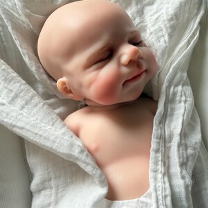 May include: A realistic newborn baby doll with smooth, light-toned skin, closed eyes, and a subtle smile. The doll is wrapped in a white, textured muslin swaddle blanket, showing only the head, shoulders, and upper chest area in a resting position.