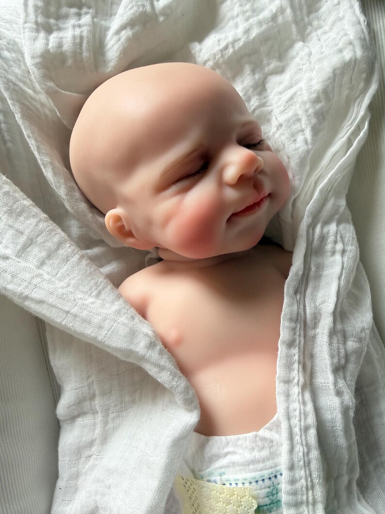 May include: A realistic newborn baby doll with smooth, light-toned skin, closed eyes, and a subtle smile. The doll is wrapped in a white, textured muslin swaddle blanket, showing only the head, shoulders, and upper chest area in a resting position.