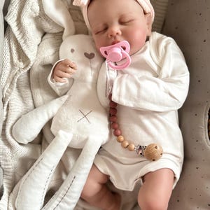 May include: A realistic baby doll with closed eyes, wearing a cream-colored long-sleeved onesie and a pale pink knotted headband. The doll has a pink pacifier attached to a wooden bead pacifier clip and is holding a white plush rabbit toy.
