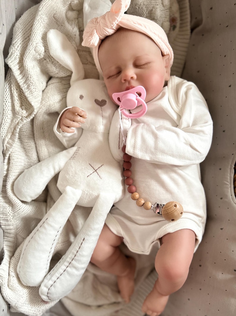 May include: A realistic baby doll with closed eyes, wearing a cream-colored long-sleeved onesie and a pale pink knotted headband. The doll has a pink pacifier attached to a wooden bead pacifier clip and is holding a white plush rabbit toy.