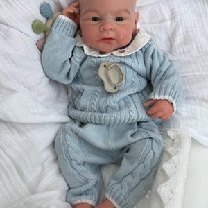 K&ouml;nnte beinhalten: Eine realistische Babypuppe mit heller Haut, bekleidet mit einem hellblauen Zopfmuster-Pullover und passender Hose mit wei&szlig;em Saum. Die Puppe tr&auml;gt ein wei&szlig;es Hemd mit Kragen darunter, einen beigen Schnuller und ein kleines, buntes Silikonspielzeug.