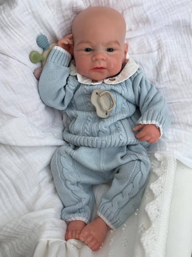 K&ouml;nnte beinhalten: Eine realistische Babypuppe mit heller Haut, bekleidet mit einem hellblauen Zopfmuster-Pullover und passender Hose mit wei&szlig;em Saum. Die Puppe tr&auml;gt ein wei&szlig;es Hemd mit Kragen darunter, einen beigen Schnuller und ein kleines, buntes Silikonspielzeug.
