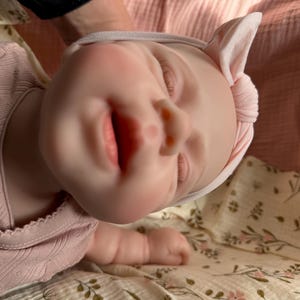 Może przedstawiać: Realistyczna lalka o jasnej karnacji, z zamkniętymi oczami i lekko otwartymi ustami. Lalka ma na sobie jasnor&oacute;żową koszulkę z długim rękawem w prążki z subtelnym wzorem oraz pasującą jasnor&oacute;żową opaskę z węzłem na głowie.