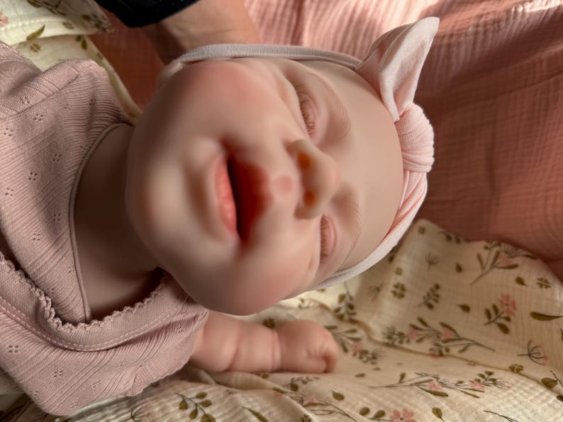 Może przedstawiać: Realistyczna lalka o jasnej karnacji, z zamkniętymi oczami i lekko otwartymi ustami. Lalka ma na sobie jasnor&oacute;żową koszulkę z długim rękawem w prążki z subtelnym wzorem oraz pasującą jasnor&oacute;żową opaskę z węzłem na głowie.