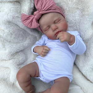 May include: A realistic newborn doll with closed eyes, wearing a plain white long-sleeved bodysuit and a dusty rose-colored ribbed fabric headband featuring a large, textured bow detail on the side, resting in a sleeping position.