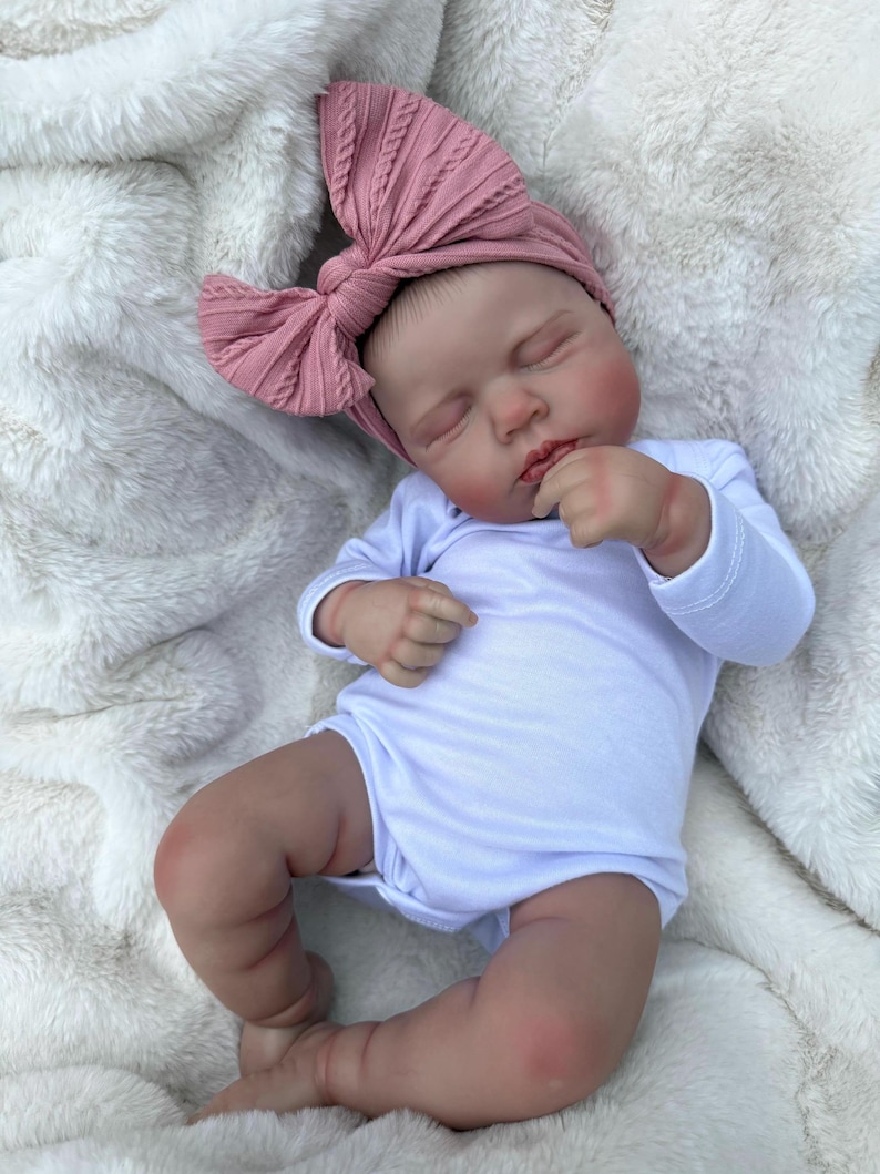May include: A realistic newborn doll with closed eyes, wearing a plain white long-sleeved bodysuit and a dusty rose-colored ribbed fabric headband featuring a large, textured bow detail on the side, resting in a sleeping position.