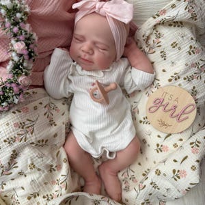 May include: A realistic newborn doll with closed eyes, wearing a white ribbed long-sleeve onesie and a light pink bow headband. The doll has a small pacifier on its chest and lies next to a wooden disc with the text It's a girl in pink script.