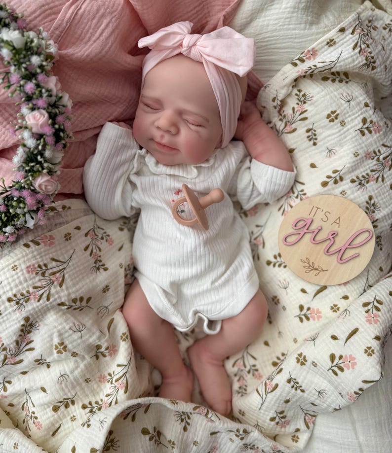 May include: A realistic newborn doll with closed eyes, wearing a white ribbed long-sleeve onesie and a light pink bow headband. The doll has a small pacifier on its chest and lies next to a wooden disc with the text It's a girl in pink script.
