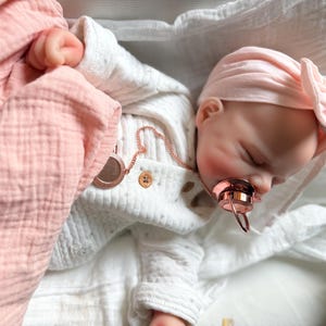 May include: A realistic baby doll with closed eyes, wearing a white long-sleeved shirt with small buttons and a light pink head wrap. The doll has a rose gold pacifier in its mouth and is wrapped in a soft, light pink muslin blanket.