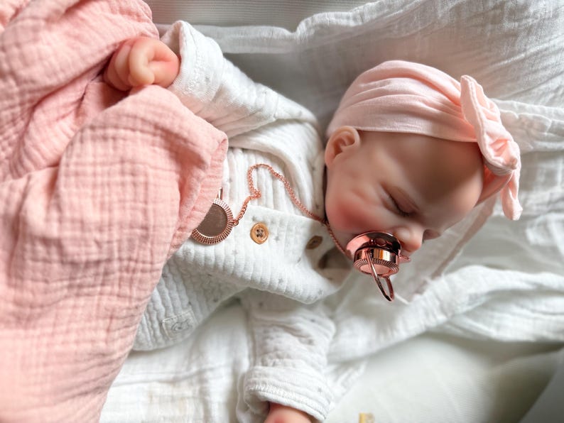 May include: A realistic baby doll with closed eyes, wearing a white long-sleeved shirt with small buttons and a light pink head wrap. The doll has a rose gold pacifier in its mouth and is wrapped in a soft, light pink muslin blanket.