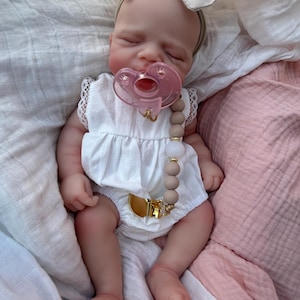 May include: A realistic baby doll with closed eyes, wearing a white sleeveless romper with lace trim and a beige fabric bow headband. The doll has a pink pacifier attached to a beaded pacifier clip featuring tan and white beads with gold accents.