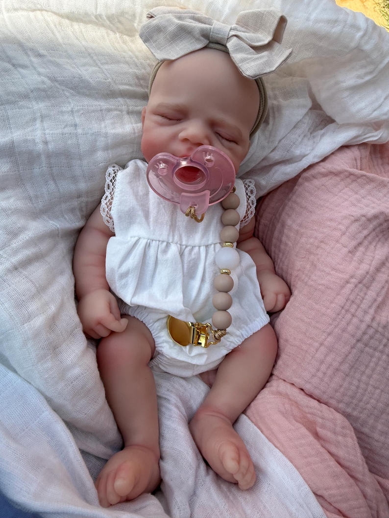 May include: A realistic baby doll with closed eyes, wearing a white sleeveless romper with lace trim and a beige fabric bow headband. The doll has a pink pacifier attached to a beaded pacifier clip featuring tan and white beads with gold accents.