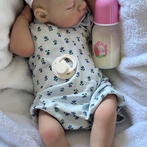 May include: A realistic baby doll with light skin, wearing a white headband with a flower and a white onesie featuring a small blue and purple floral pattern. The doll has a cream-colored pacifier on its chest and is next to a baby bottle with a pink cap and a graphic of a white rabbit with a strawberry.
