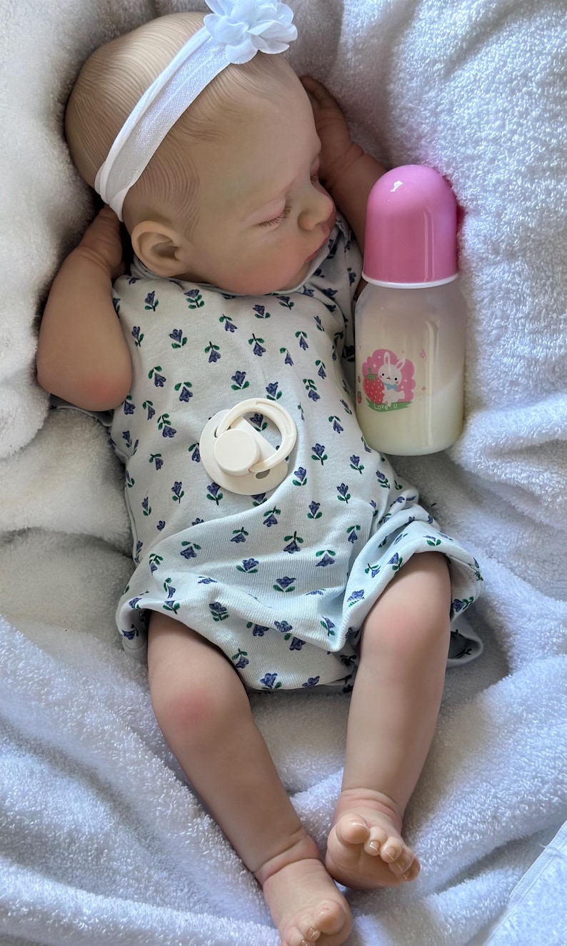 May include: A realistic baby doll with light skin, wearing a white headband with a flower and a white onesie featuring a small blue and purple floral pattern. The doll has a cream-colored pacifier on its chest and is next to a baby bottle with a pink cap and a graphic of a white rabbit with a strawberry.