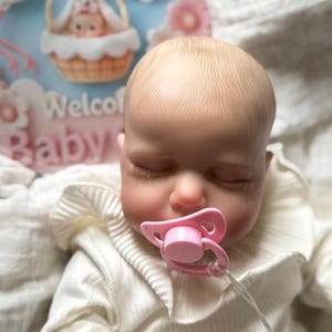 May include: A realistic baby doll with a pale complexion and closed eyes, wearing a cream-coloured ribbed long-sleeve shirt with a ruffled collar. The doll has a pink pacifier in its mouth and is positioned in front of a sign that says Welcome Baby.
