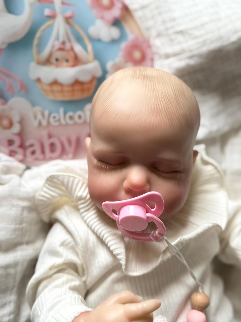 May include: A realistic baby doll with a pale complexion and closed eyes, wearing a cream-coloured ribbed long-sleeve shirt with a ruffled collar. The doll has a pink pacifier in its mouth and is positioned in front of a sign that says Welcome Baby.