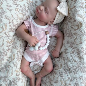 May include: A sleeping baby doll wearing a pink and white striped onesie and a cream-colored bow headband. The doll is holding a pacifier with white beads and wooden accents. The baby is lying on a cream-colored blanket with a woodland animal print.