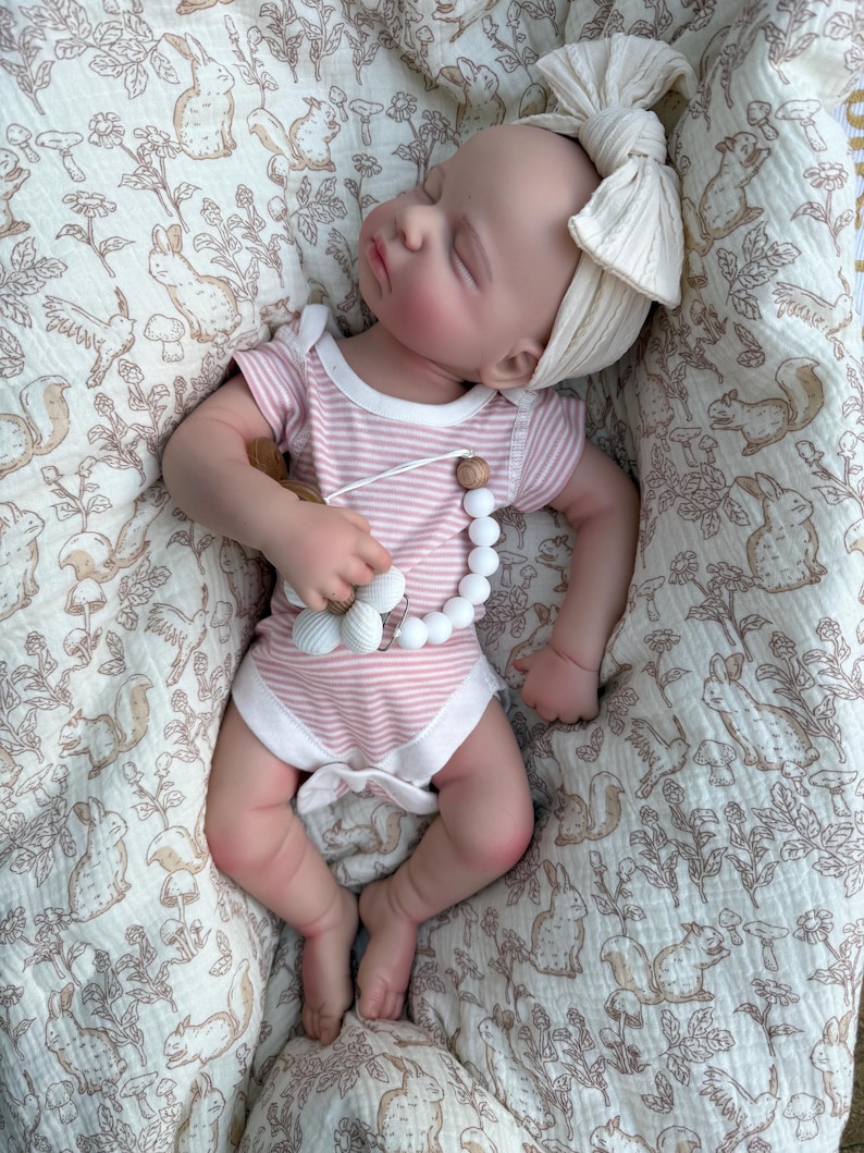 May include: A sleeping baby doll wearing a pink and white striped onesie and a cream-colored bow headband. The doll is holding a pacifier with white beads and wooden accents. The baby is lying on a cream-colored blanket with a woodland animal print.