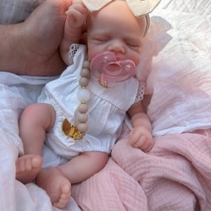 May include: A realistic baby doll with closed eyes, wearing a white sleeveless romper with lace trim and a beige fabric bow headband. The doll has a translucent pink pacifier and a pacifier clip featuring light brown and white beads.