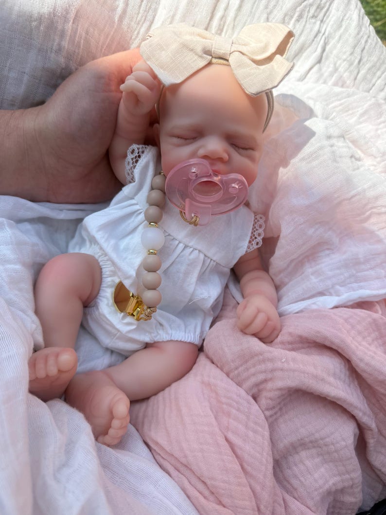 May include: A realistic baby doll with closed eyes, wearing a white sleeveless romper with lace trim and a beige fabric bow headband. The doll has a translucent pink pacifier and a pacifier clip featuring light brown and white beads.