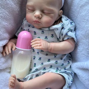 May include: A realistic baby doll with closed eyes, wearing a white short-sleeved romper featuring a small blue and green floral pattern and a white headband with a flower. The doll is holding a plastic baby bottle with a pink cap.
