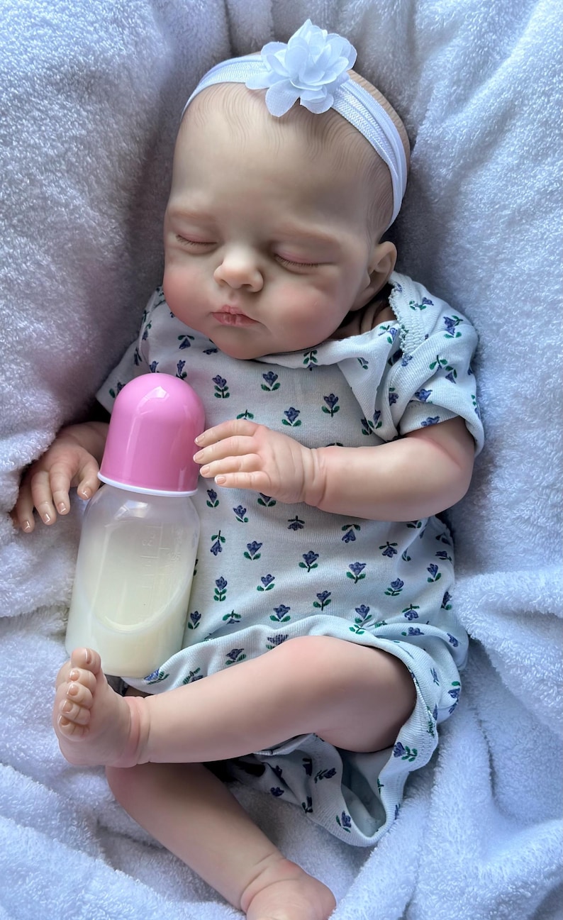 May include: A realistic baby doll with closed eyes, wearing a white short-sleeved romper featuring a small blue and green floral pattern and a white headband with a flower. The doll is holding a plastic baby bottle with a pink cap.