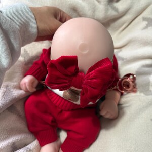 Pode incluir: Uma boneca careca com cabe&ccedil;a lisa e clara usando um grande la&ccedil;o de veludo vermelho texturizado. A boneca est&aacute; vestida com um su&eacute;ter vermelho de manga comprida e cal&ccedil;as vermelhas combinando com punhos canelados. Uma chupeta ouro rosa est&aacute; presa.