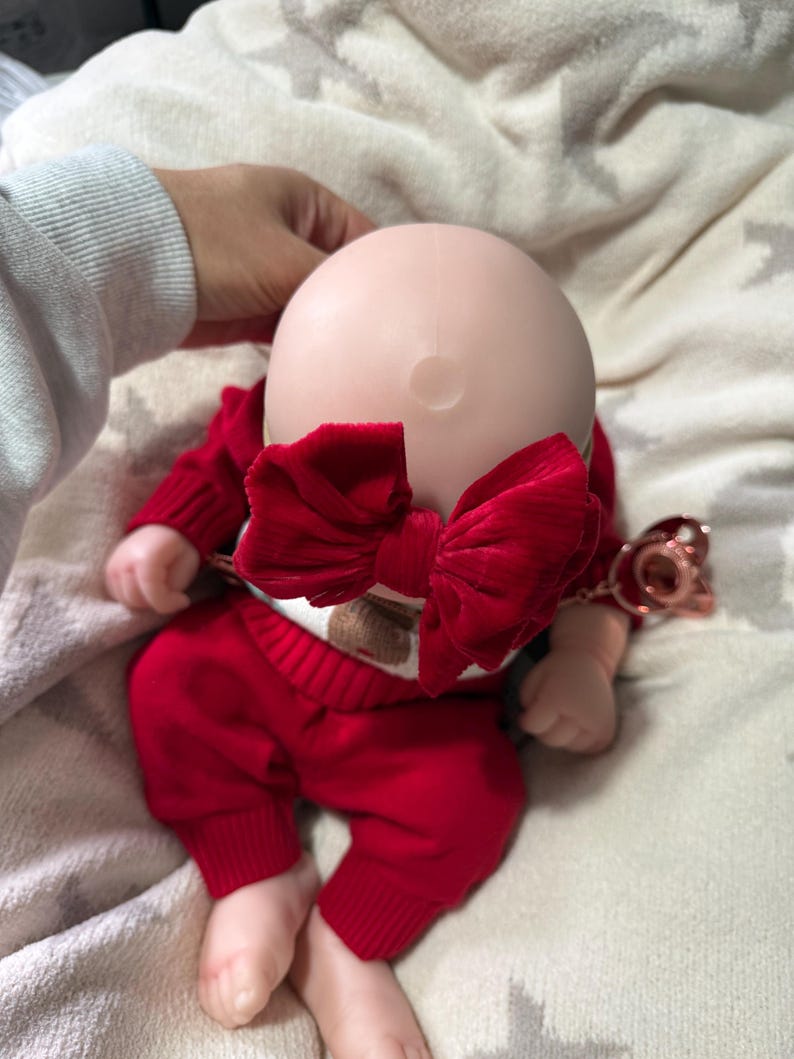 Pode incluir: Uma boneca careca com cabe&ccedil;a lisa e clara usando um grande la&ccedil;o de veludo vermelho texturizado. A boneca est&aacute; vestida com um su&eacute;ter vermelho de manga comprida e cal&ccedil;as vermelhas combinando com punhos canelados. Uma chupeta ouro rosa est&aacute; presa.