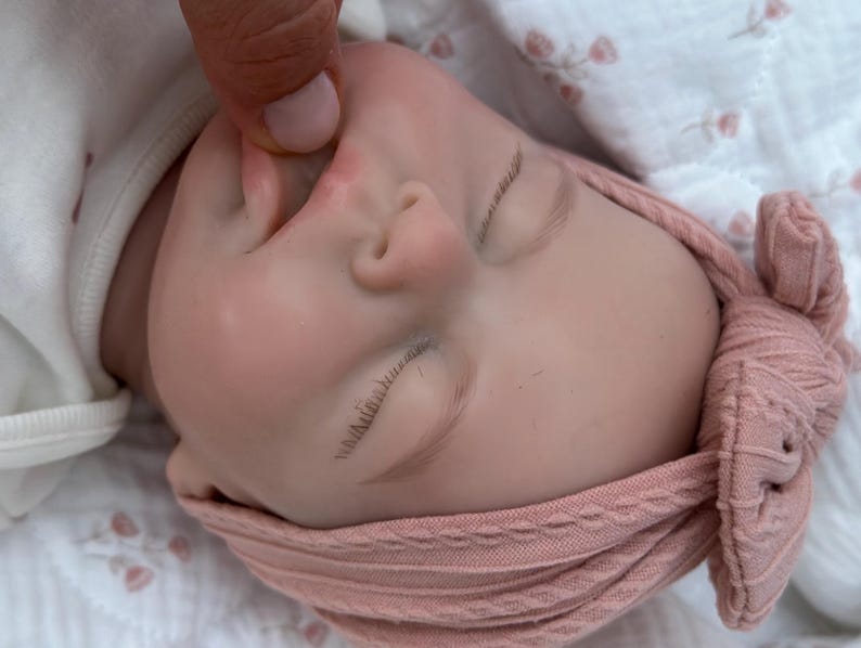 Może przedstawiać: Realistyczna lalka dla dziecka z zamkniętymi oczami, delikatnymi rzęsami i miękką, r&oacute;żową cerą. Lalka ma na sobie pasującą r&oacute;żową dzianinową opaskę na głowę. Lalka leży na białej powierzchni.