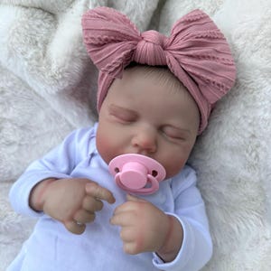 May include: A realistic newborn doll with closed eyes, wearing a white long-sleeved onesie and a dusty rose-colored fabric headband featuring a large, textured bow. The doll has a light pink pacifier in its mouth and is positioned with its hands near its face.