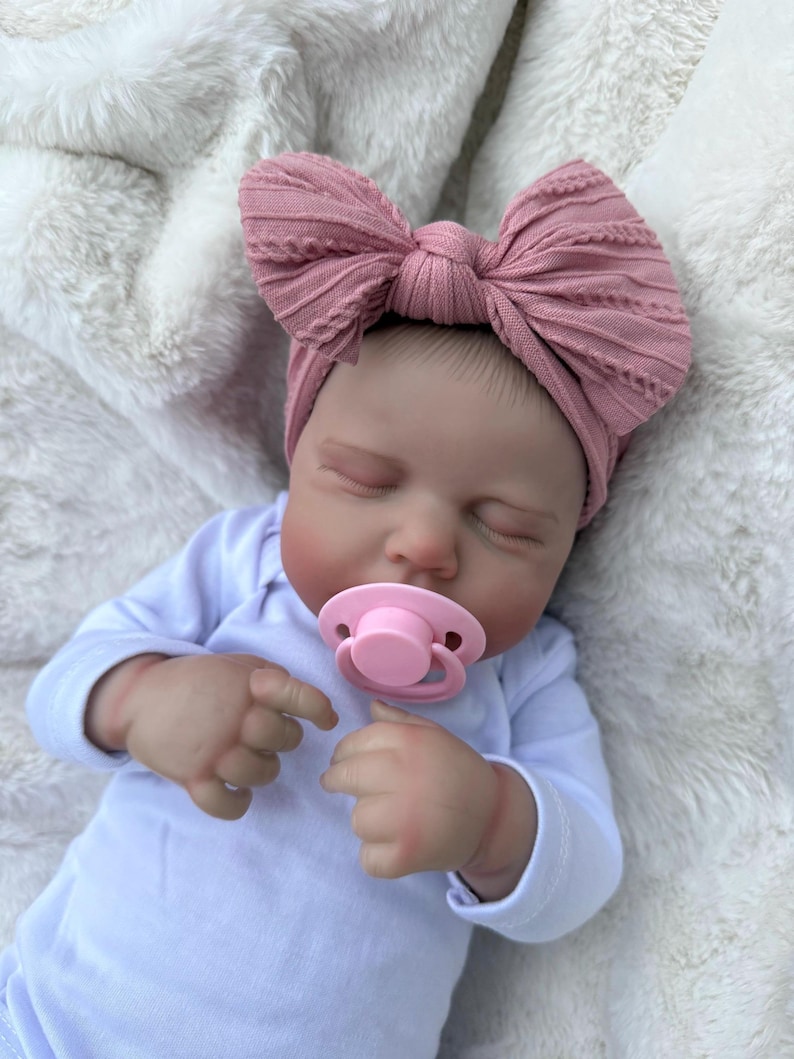 May include: A realistic newborn doll with closed eyes, wearing a white long-sleeved onesie and a dusty rose-colored fabric headband featuring a large, textured bow. The doll has a light pink pacifier in its mouth and is positioned with its hands near its face.