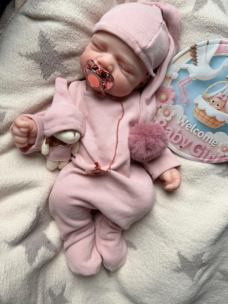 Pu&ograve; includere: Una bambola realistica che indossa una tutina a maniche lunghe con piedini a coste rosa chiaro e un cappellino abbinato. La bambola ha un ciuccio color oro rosa con catenella abbinata e un piccolo orsetto di peluche. Un cartello circolare recita Welcome Baby Girl.
